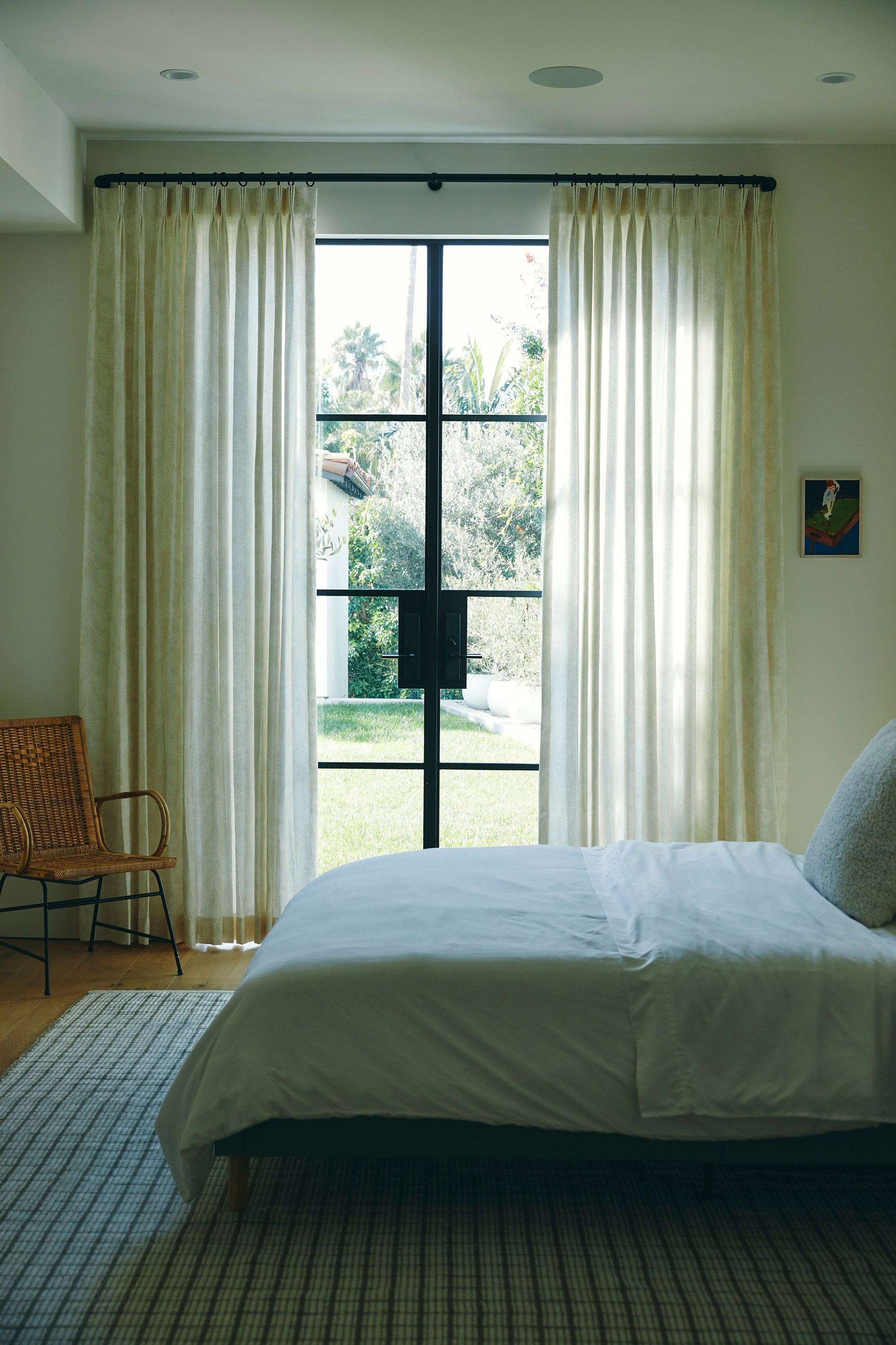 Bright bedroom with white bedding, beige sheer curtains, large window, wicker chair, gray rug, garden view.