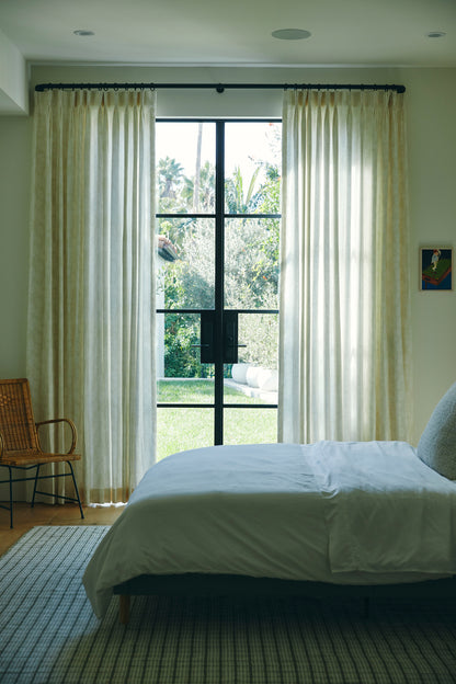 Bright bedroom with a 9' x 12' rug on the floor, white bed, sheer cream curtains, a black-framed glass door to a garden, & a wicker chair beside the bed.