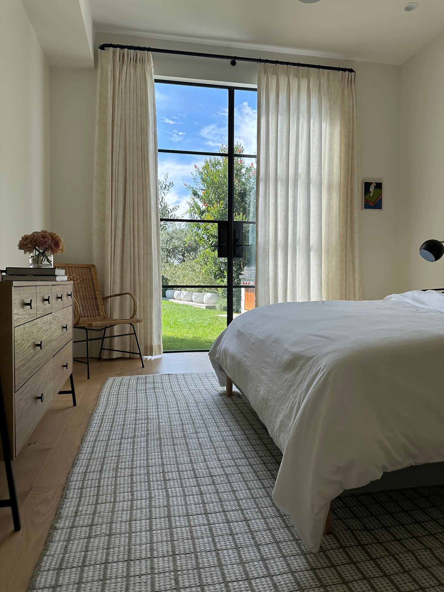 Bright bedroom with white bedding, light wood dresser, wicker chair, beige curtains, and a glass door to a green garden.