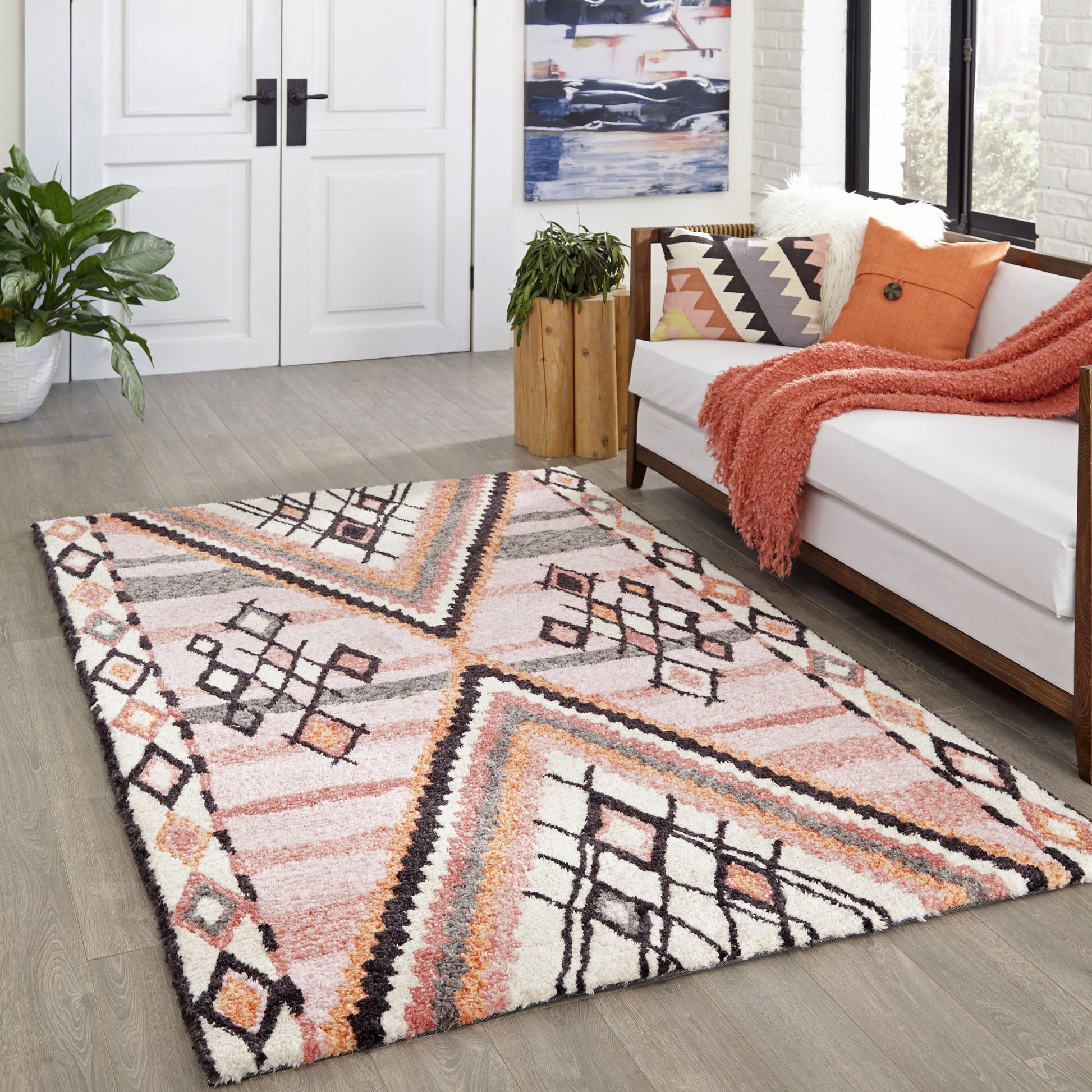 Shaggy geometric area rug in pink, peach, cream with black accents on light wood floor beside white sofa and orange throw.