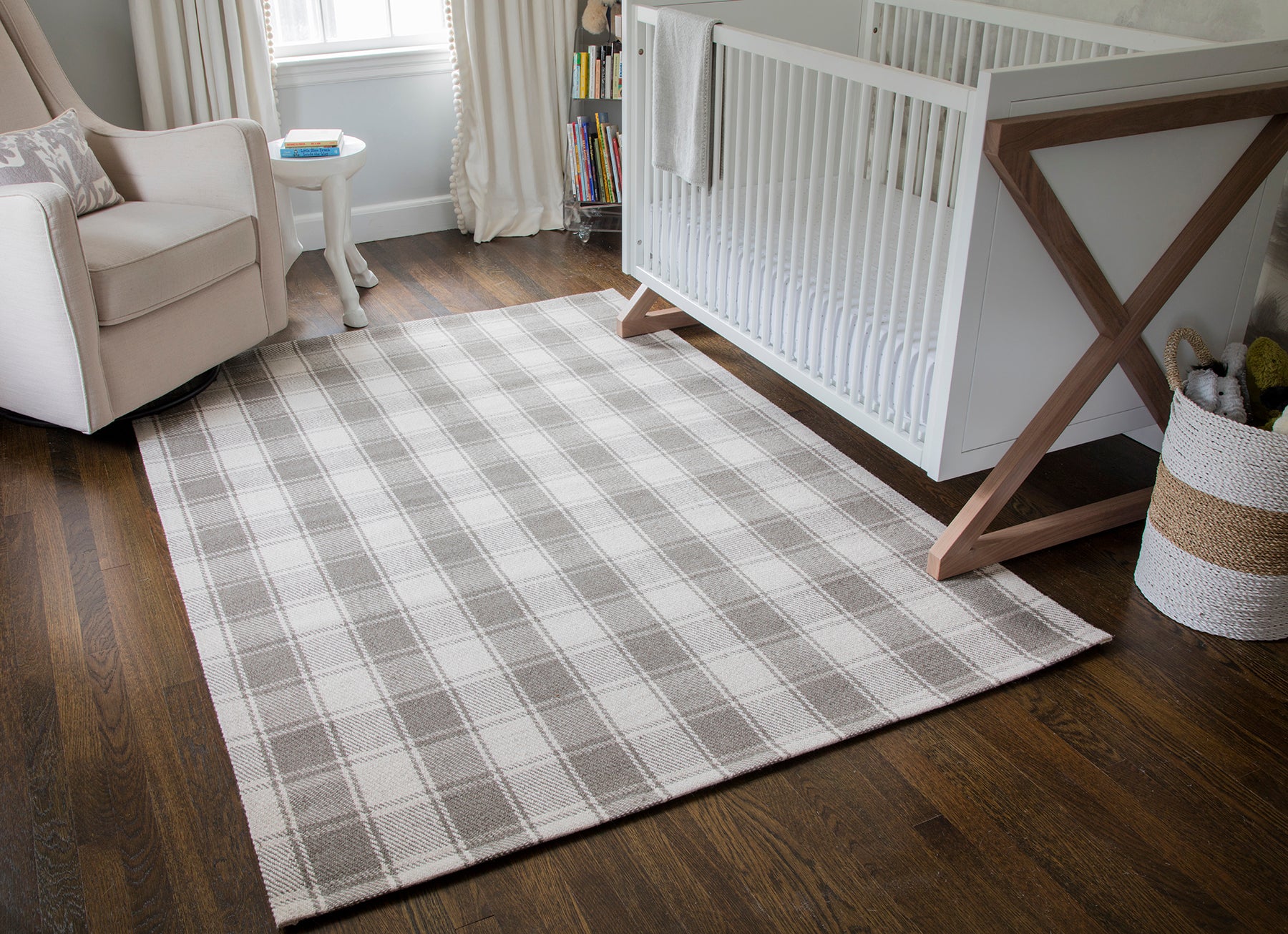 Gray and ivory plaid area rug on dark hardwood floor beside a white crib; beige armchair and woven basket in a nursery.