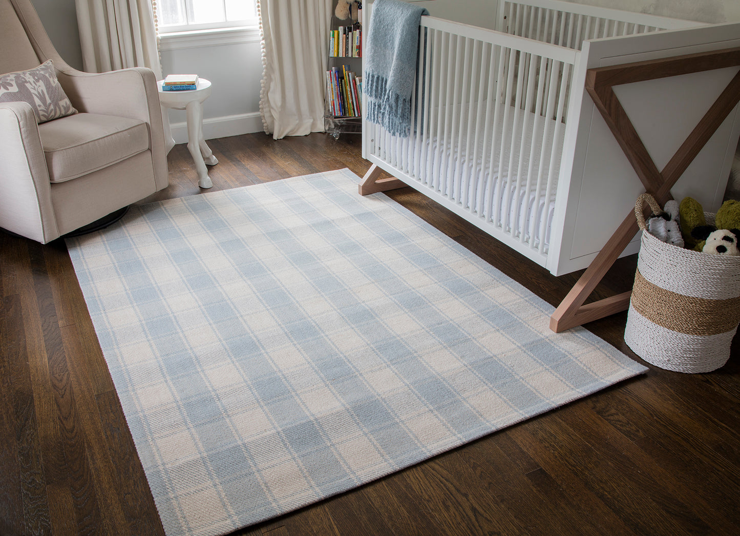Blue and white plaid area rug in nursery with a white crib and beige armchair on dark wood floor.