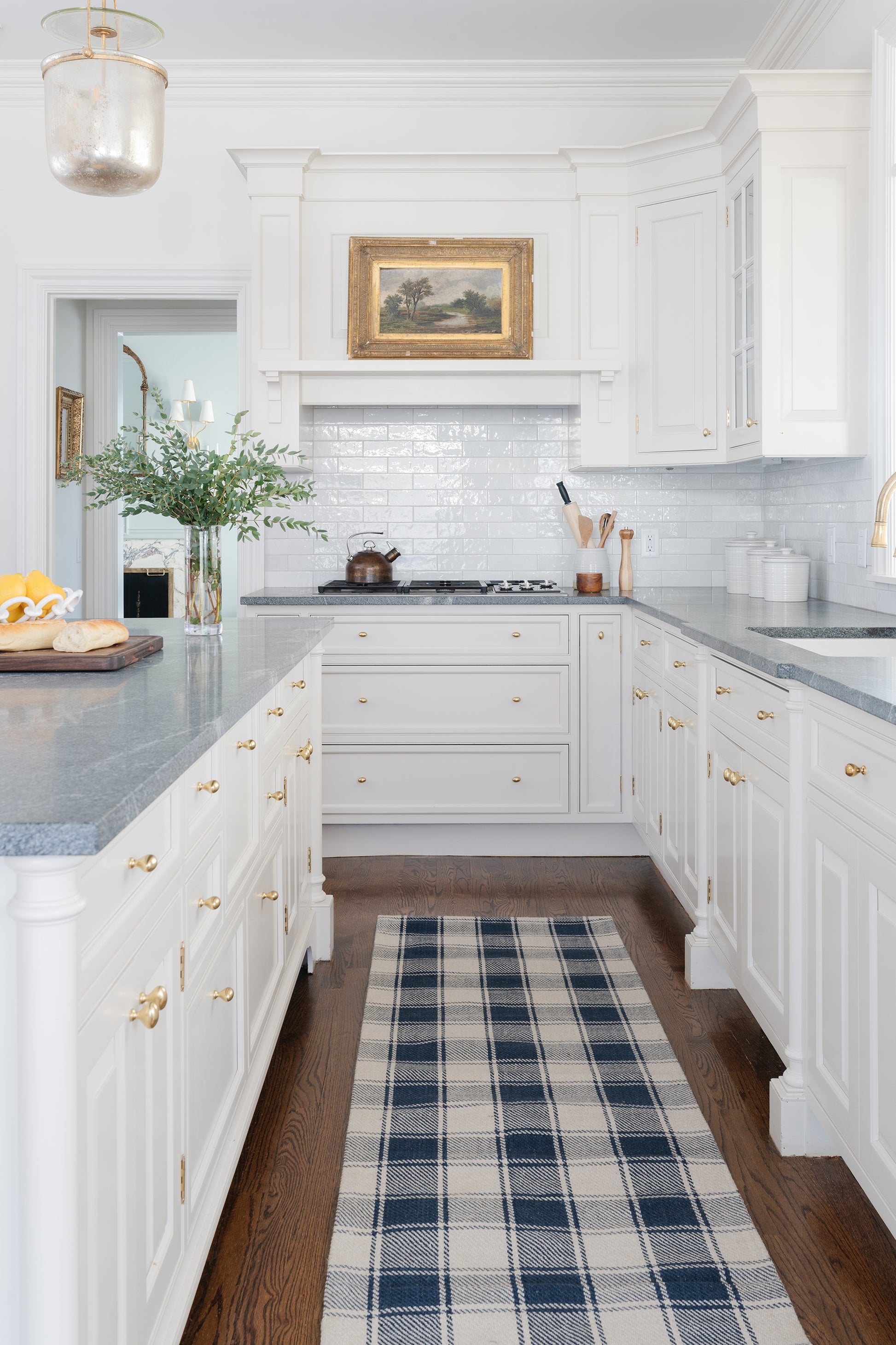 Bright white kitchen with gray countertops, gold hardware, subway tile backsplash, landscape painting, navy plaid runner.