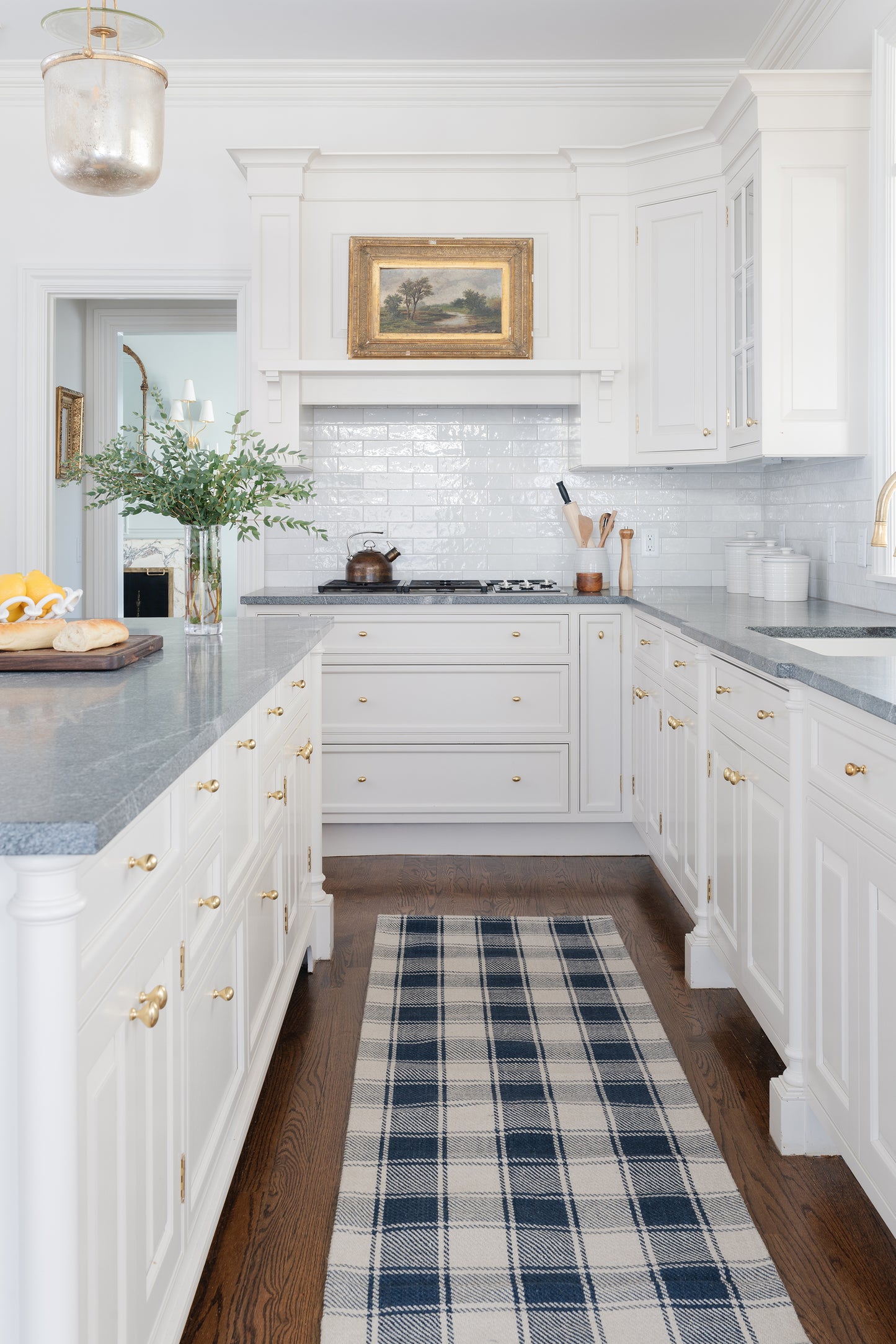 Bright white kitchen with gray countertops, brass hardware, plaid rug, copper kettle on stove, framed landscape above range.