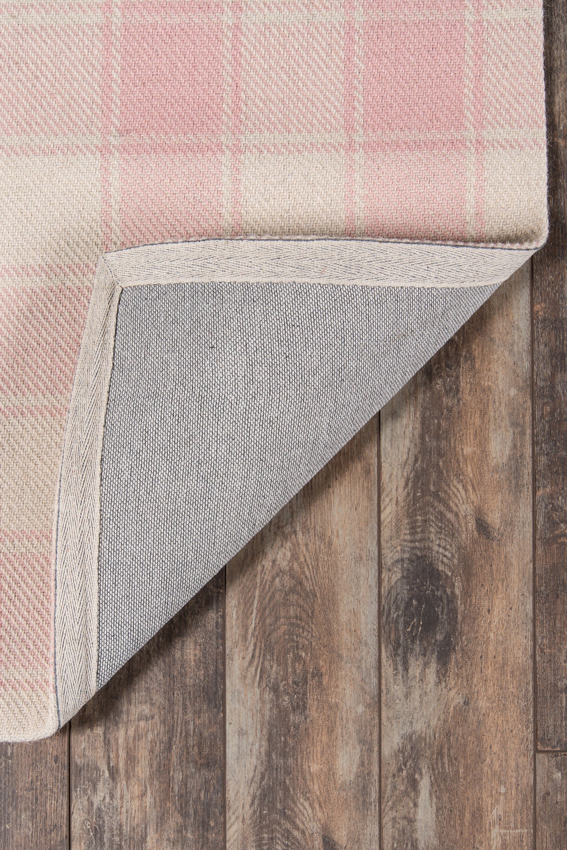 Rectangular pink plaid rug with beige border, woven texture, corner folded to reveal grey backing on wooden floor.