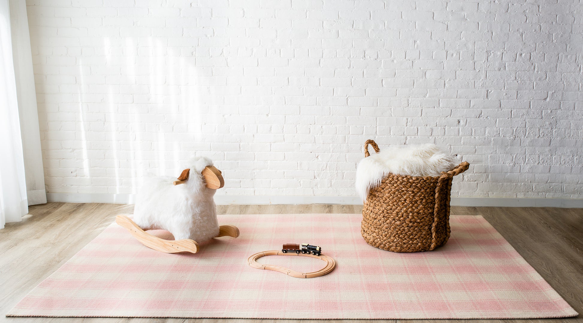 Soft pink plaid rug with a white plush rocking sheep, wooden train on tracks, and a woven basket with faux fur throw.