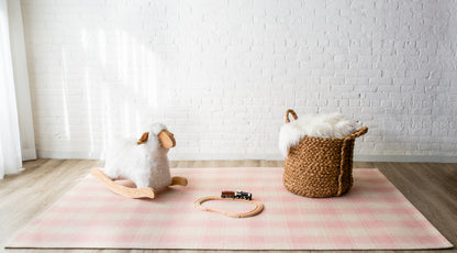 Soft pink plaid rug with a white plush rocking sheep, wooden train on tracks, and a woven basket with faux fur throw.