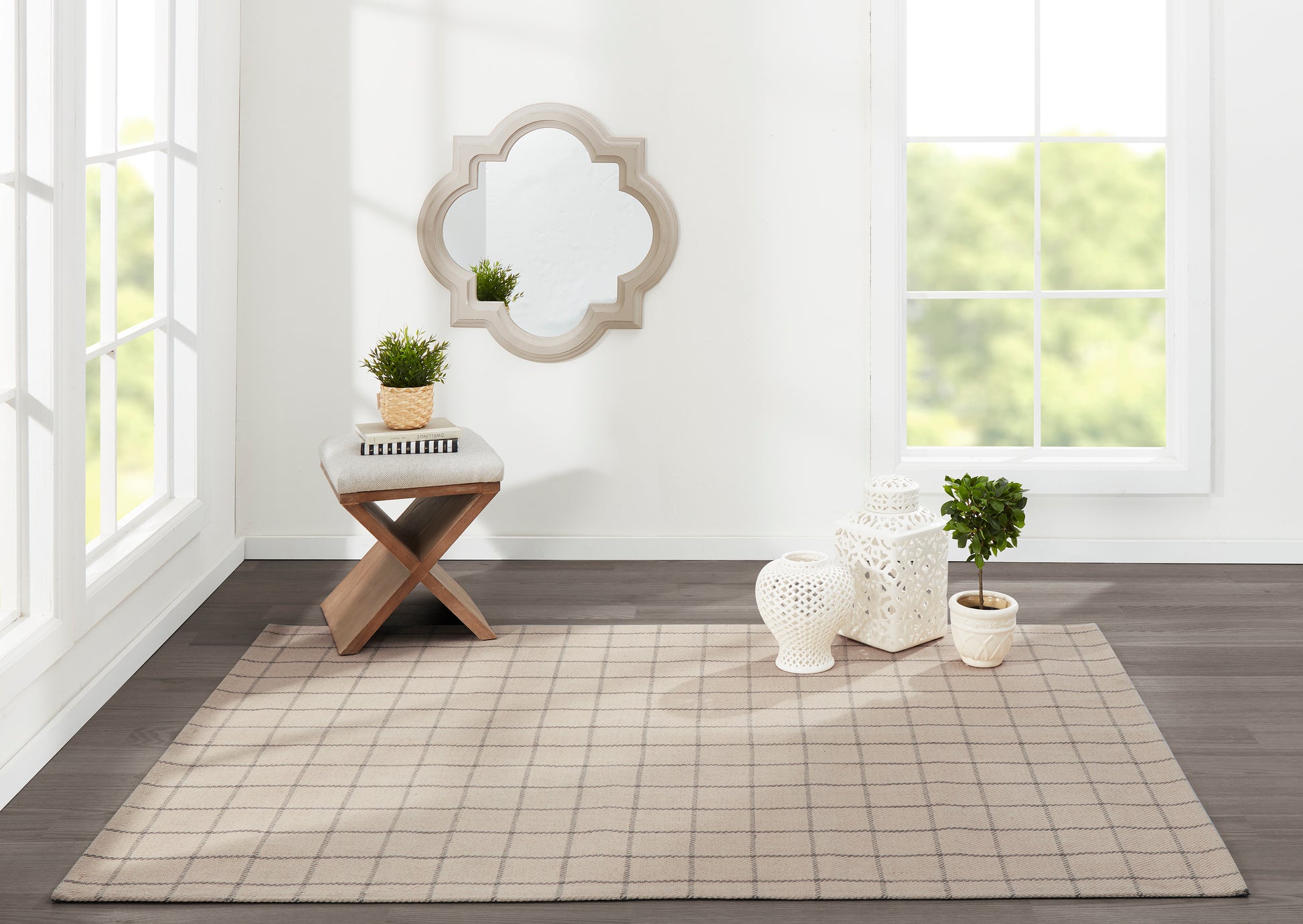 Beige grid rug on dark wood floor with wooden X-stool, potted plant and books, quatrefoil wall mirror, white lanterns.