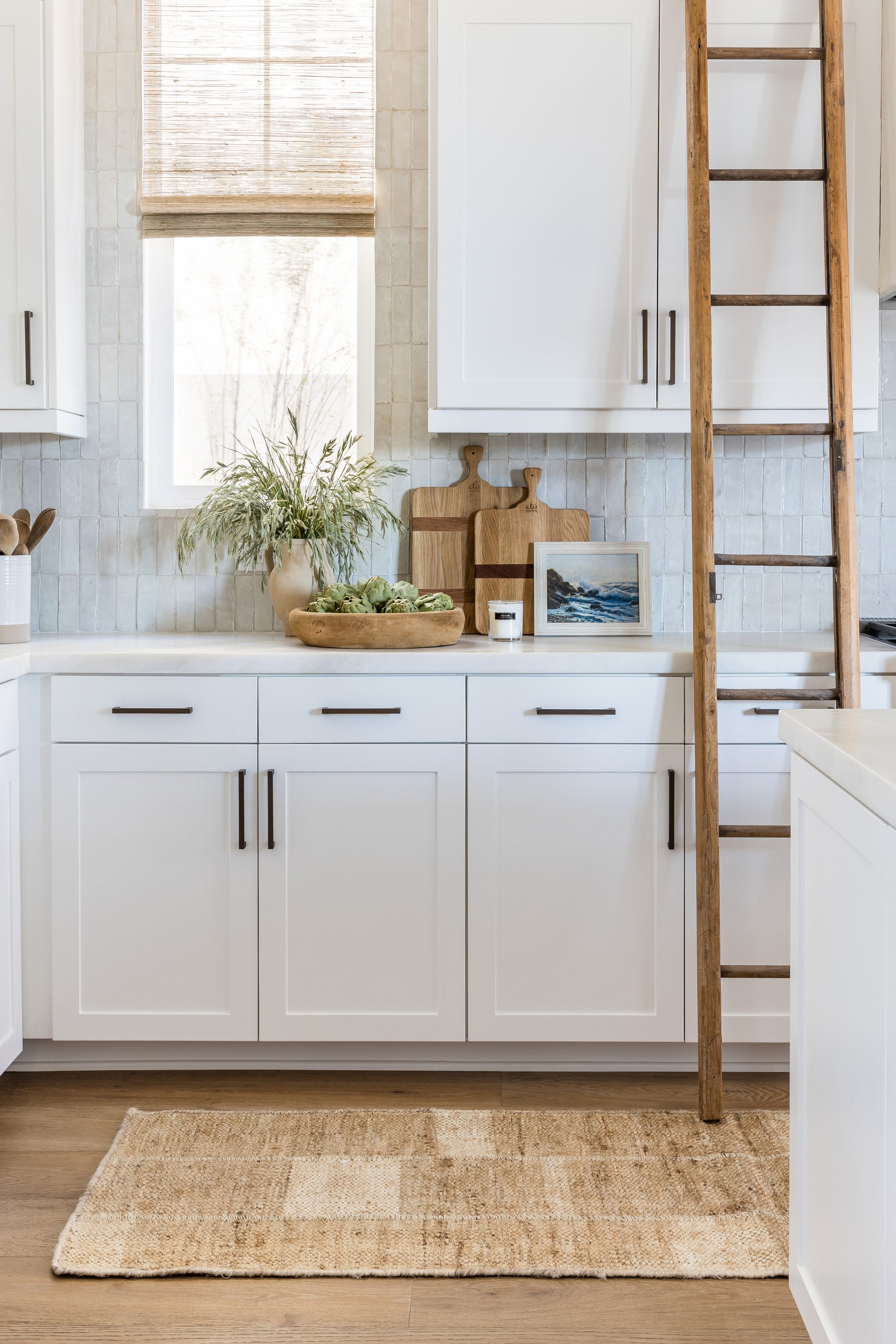 Bright white kitchen with wood countertop, natural jute rug, wooden ladder, potted plant, cutting boards, framed ocean photo.