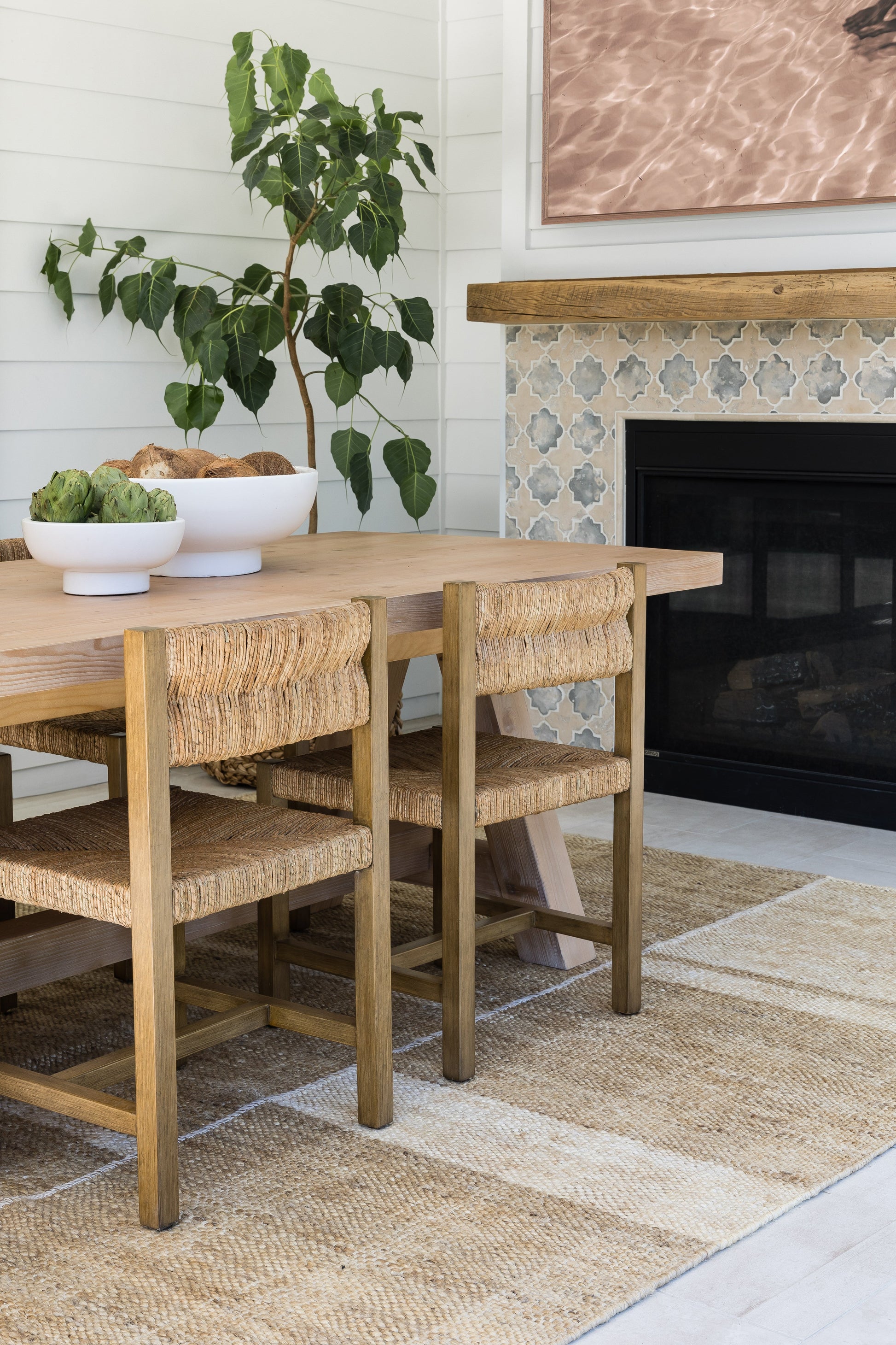 Rustic wooden dining table with woven rattan chairs, large potted plant, white bowls of artichokes, jute rug, tiled fireplace.