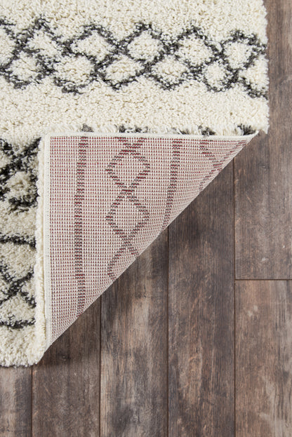 Maya Ivy ivory shag rug with black geometric lattice, underside shows pink-beige weave, on dark wood floor.