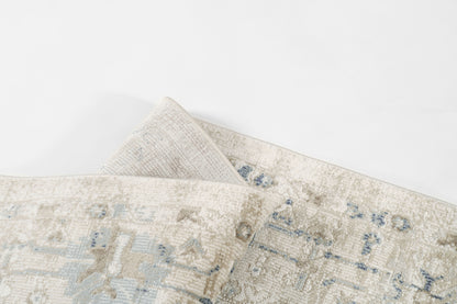 Cream-beige rug with pale blue faded pattern, woven texture, folded corner.