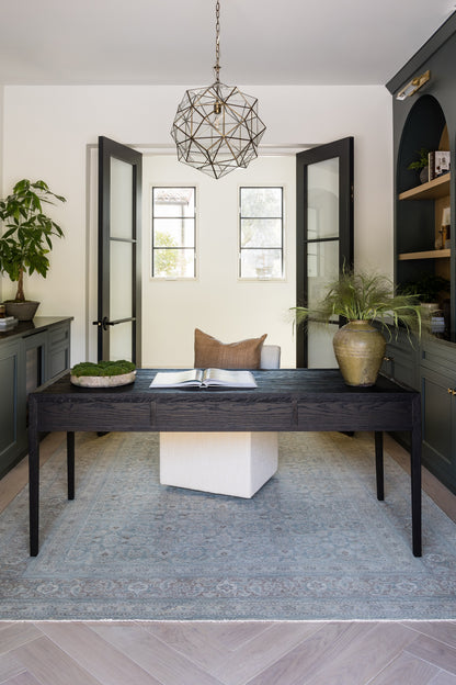 Dark wood desk with white upholstered stool, blue patterned rug, potted plant, open book, and geometric metal pendant light.