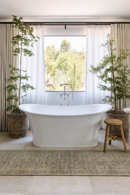 White freestanding bathtub with chrome faucet, in a bright bathroom with potted trees, beige rug, and wooden stool.