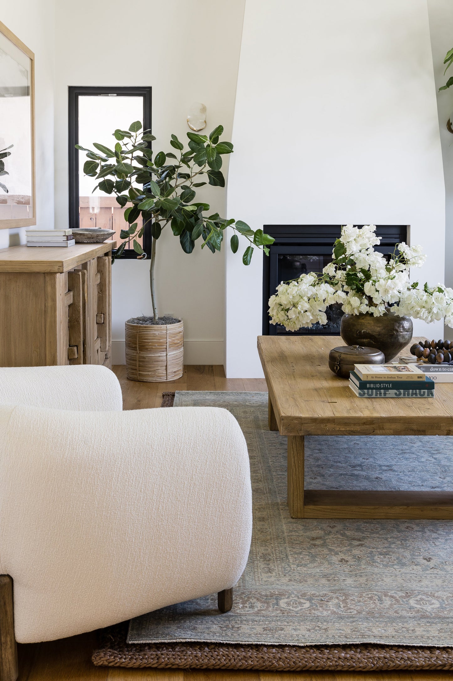 Bright modern living room with white boucle sofa, natural wood coffee table, fiddle-leaf fig in woven planter, white flowers.
