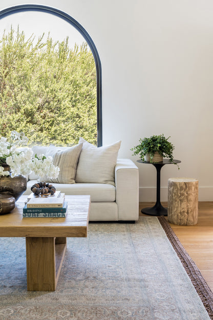 Bright living room with arched window, cream sofa, wooden coffee table, white flowers, and greenery on a pale rug