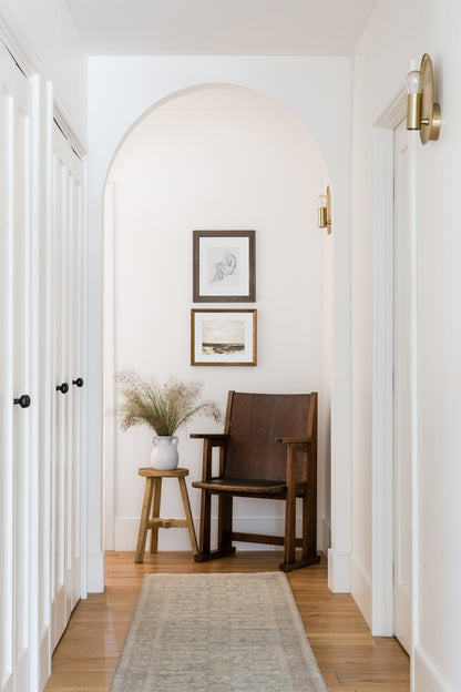 9' x 12' rug in an arched white hallway scene with a dark wood chair, a small stool with a vase of dried grasses, framed art, and brass wall sconces.
