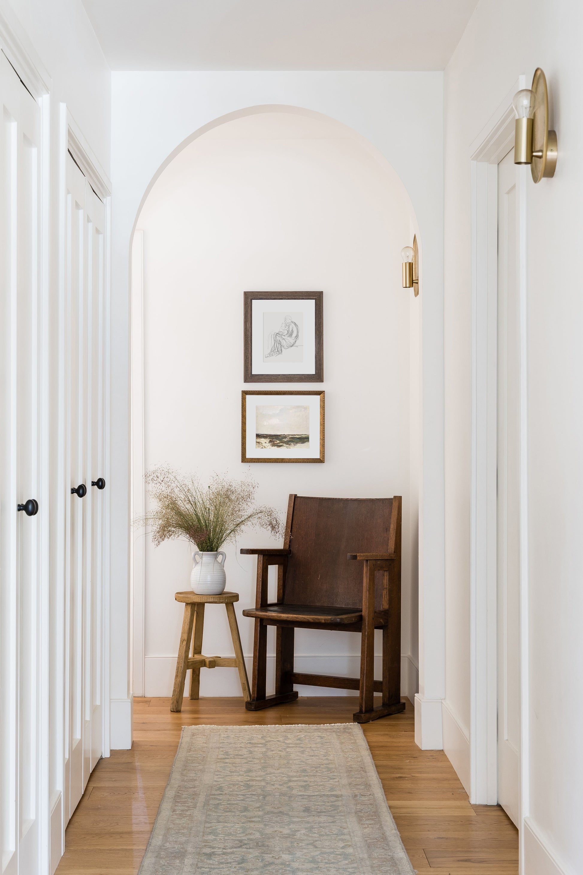 White hallway arch, dark wood armchair, small stool with vase of dried grasses, framed prints, brass sconces, pale rug