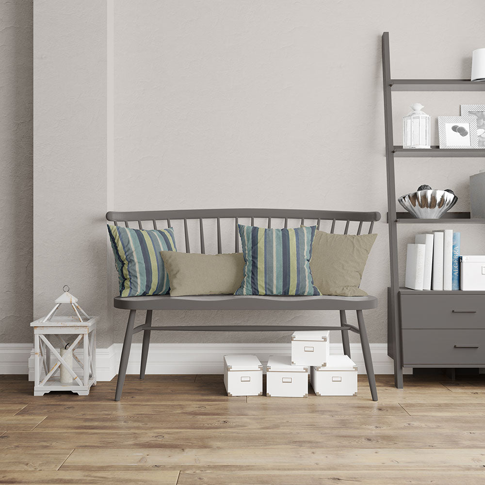 Grey wooden bench with a slatted back, striped teal-blue cushions and beige pillows, beside a grey shelving unit on a wood floor.