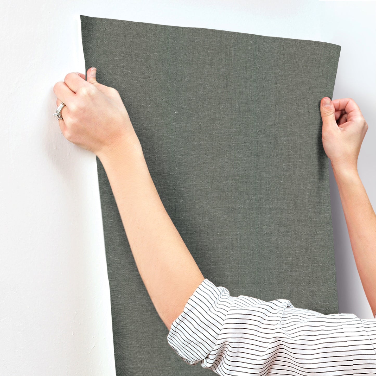 Hands hold a gray textured wallpaper swatch, rectangular and ready to mount on a white wall.