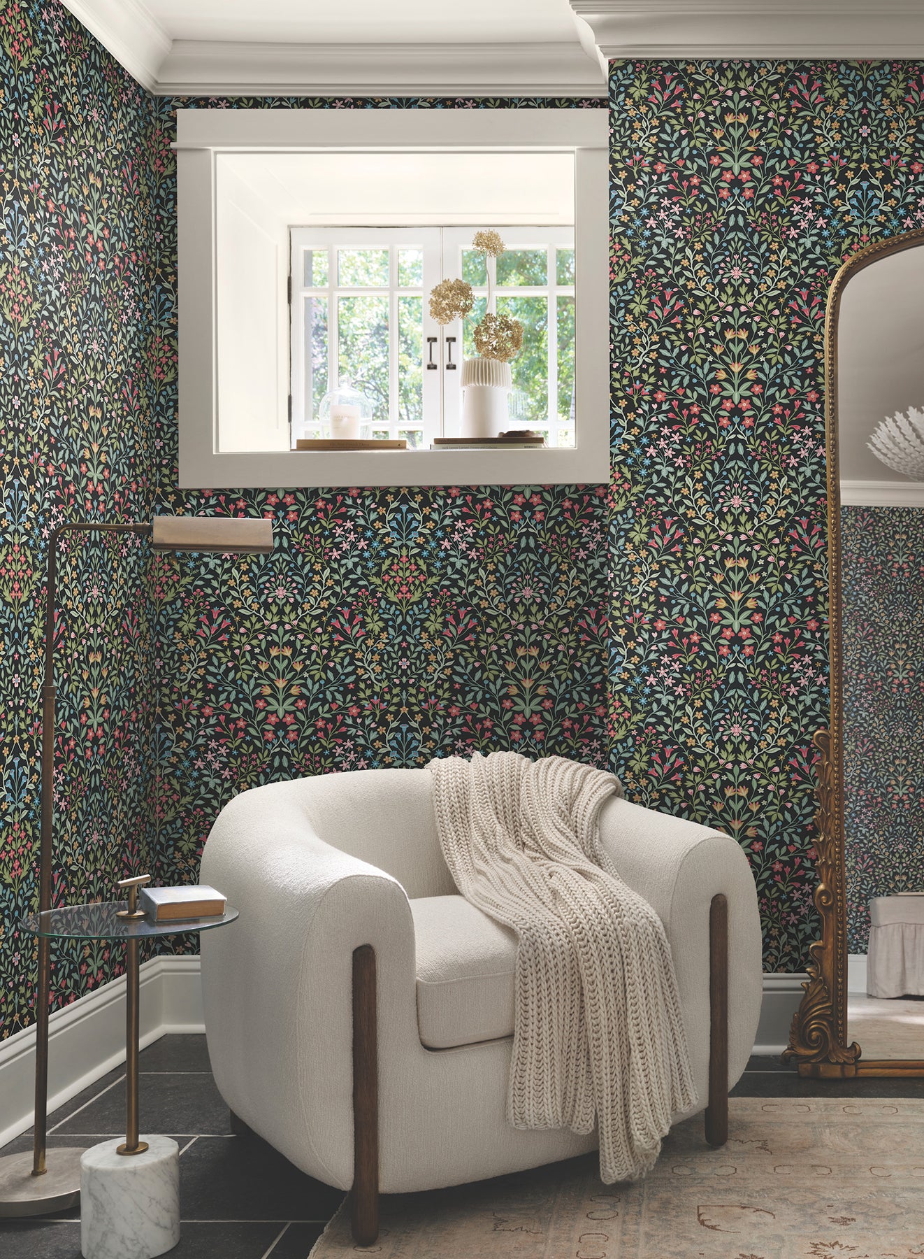 Room with black and multicolor botanical wallpaper, cream curved armchair with knit throw, window light, gold floor lamp, marble side table and standing mirror.