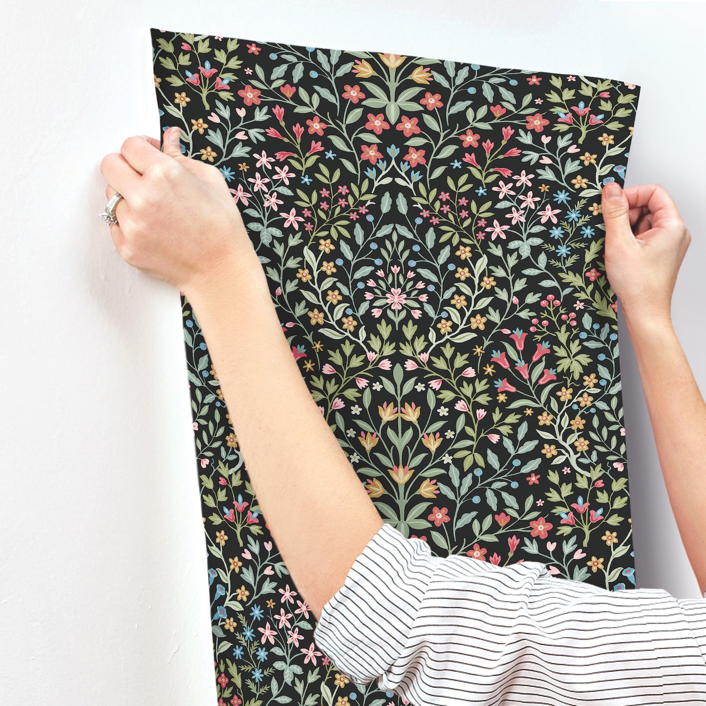 Black wallpaper swatch with a vibrant botanical pattern of multicolored leaves and blooms, held by two hands.