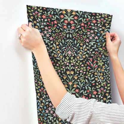 Black wallpaper swatch with a vibrant botanical pattern of multicolored leaves and blooms, held by two hands.
