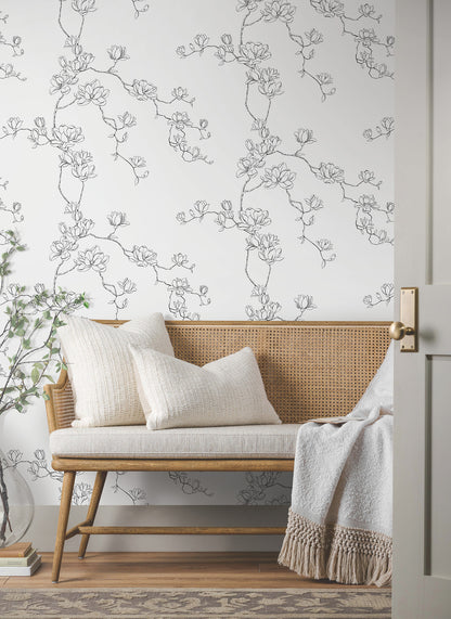 Room with black botanical branches wallpaper on the wall; a rattan bench with cream cushions is visible.
