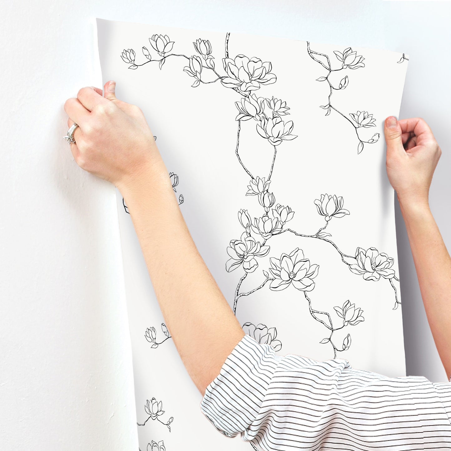 White wallpaper sheet with black magnolia branch line art, held by hands in striped sleeves.