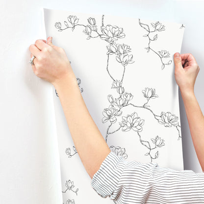 White wallpaper sheet with black magnolia branch line art, held by hands in striped sleeves.