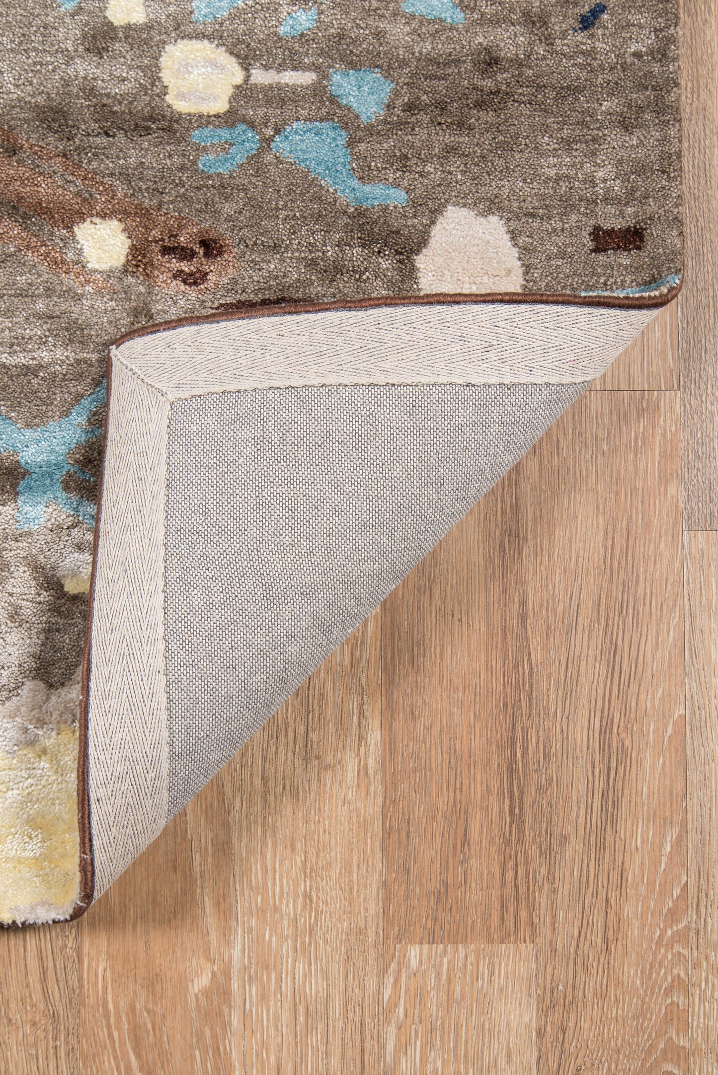 Brown-gray patterned area rug with blue, teal and yellow accents; beige herringbone underside; brown edge trim.