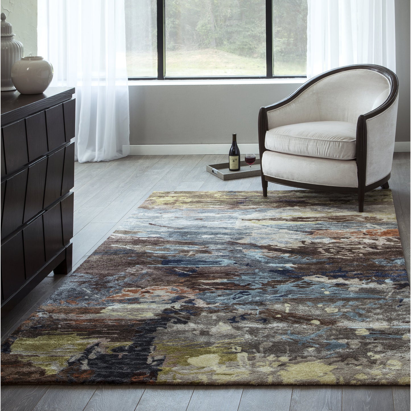 Living room with multicolor abstract rug, cream upholstered armchair with dark wood frame, tray with wine by window.