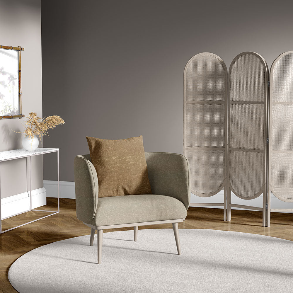 Beige fabric armchair with light wood legs and brown cushion, on circular beige rug; rattan folding screen backdrop.