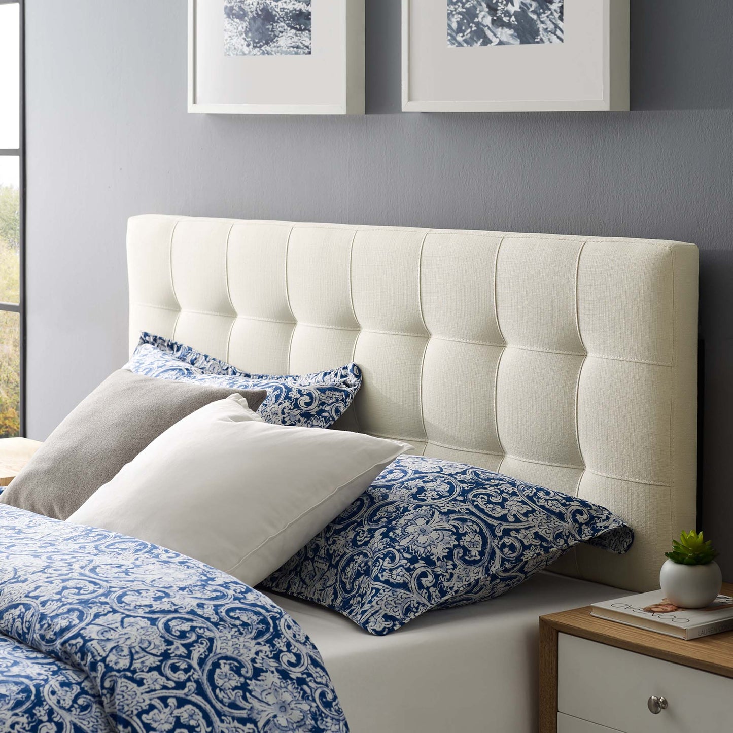 White tufted upholstered headboard on a bed with blue paisley bedding, neutral pillows, gray wall, and a wooden nightstand with a plant.