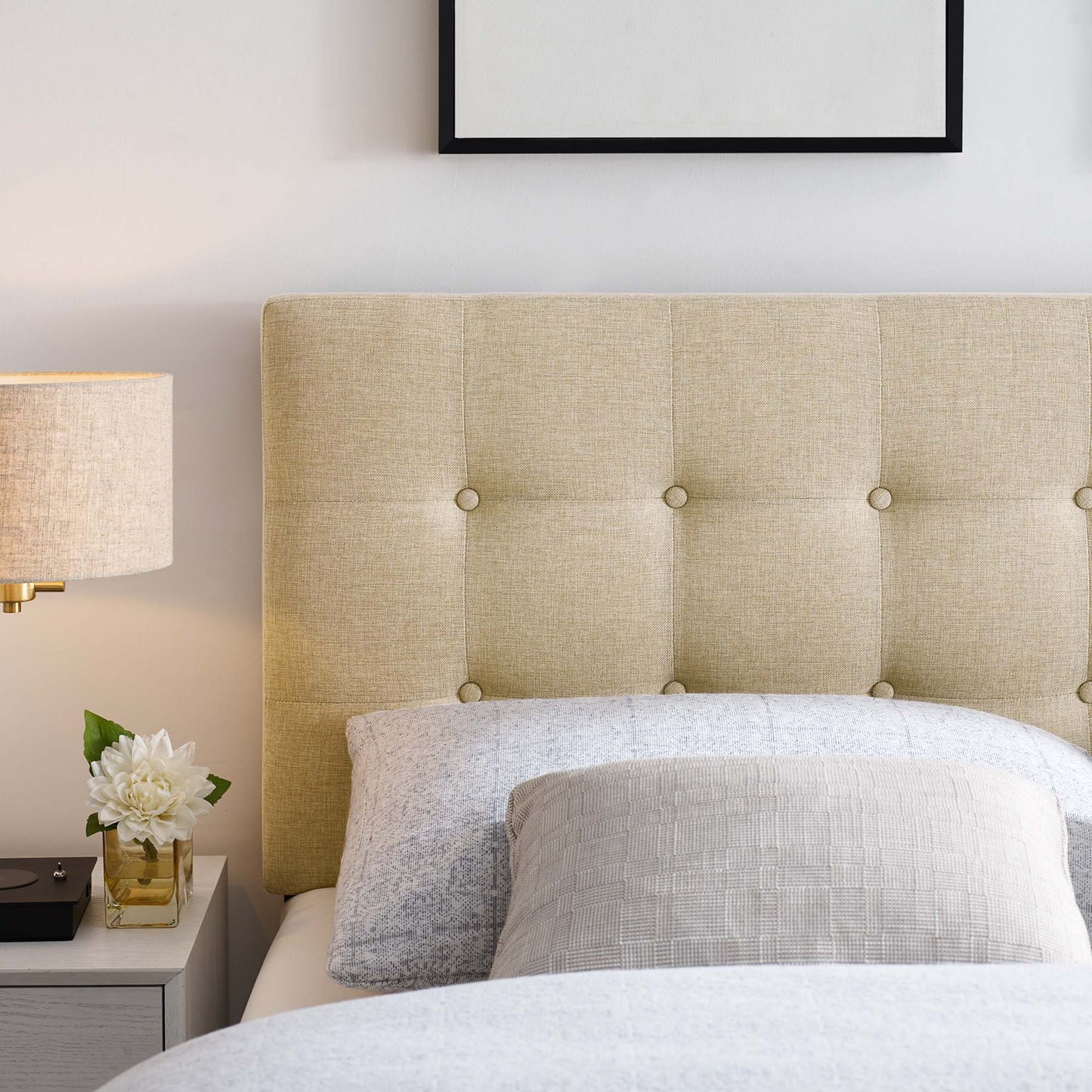 Brown tufted upholstered headboard with button details, light gray bedding, & bedside table with lamp and white flowers.
