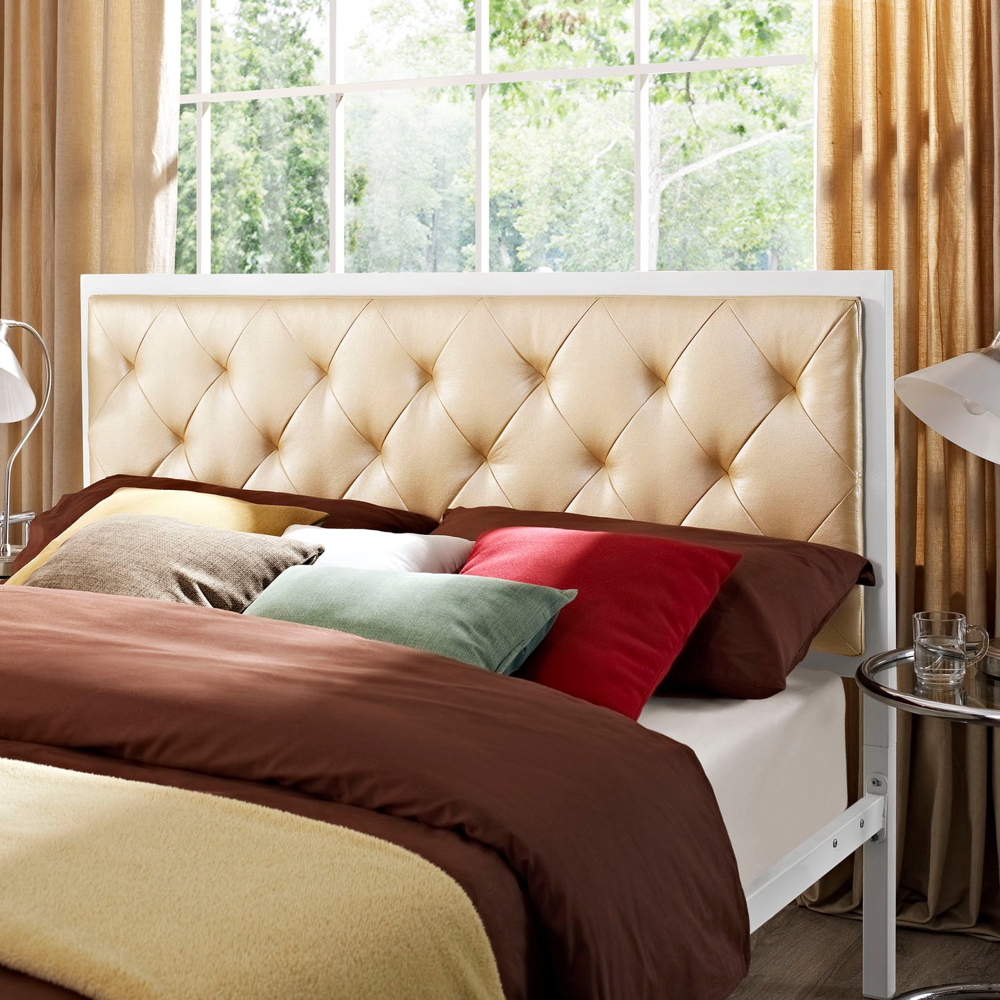 Beige tufted headboard bed with white frame, brown duvet, and multicolor throw pillows in a sunlit bedroom.