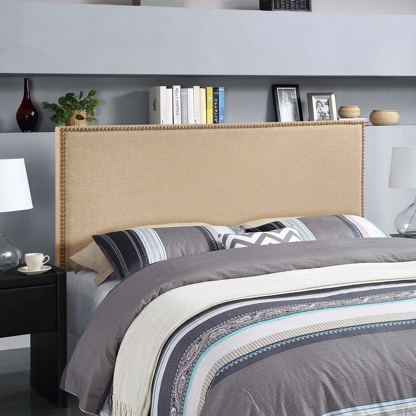 Brown upholstered queen headboard with nailhead trim; gray striped bedding; shelves with books and lamps.