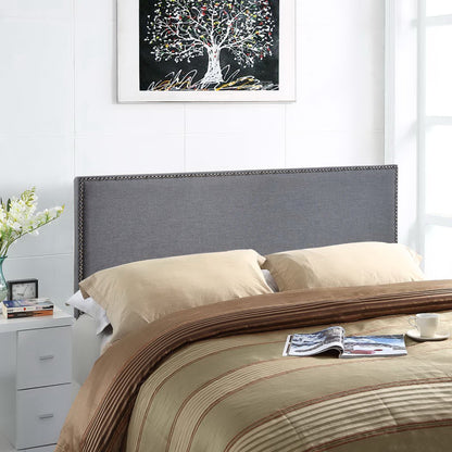 Grey upholstered bed headboard with nailhead trim, beige sheets, brown striped comforter; nightstand, vase of flowers, & tree artwork above.