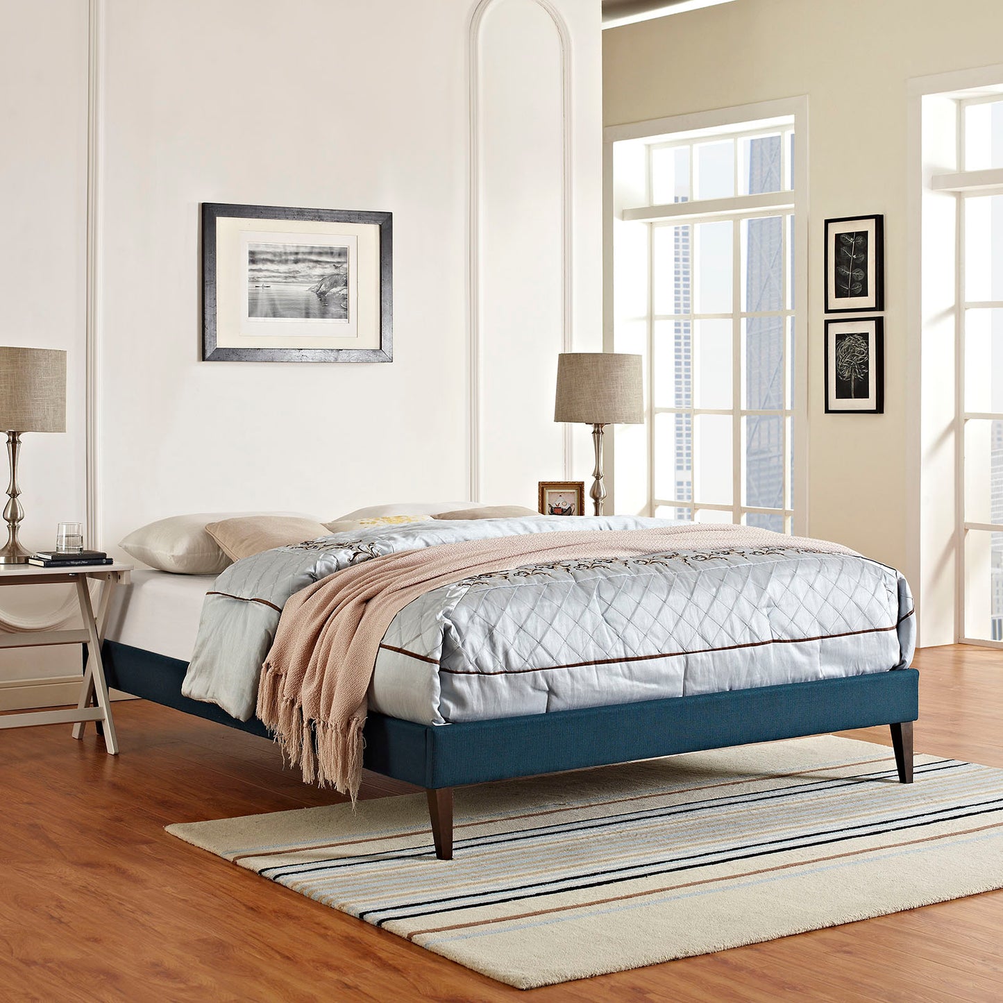 Azure upholstered platform bed with quilted gray comforter, brown piping, beige throw; dark wood legs in a modern bedroom.