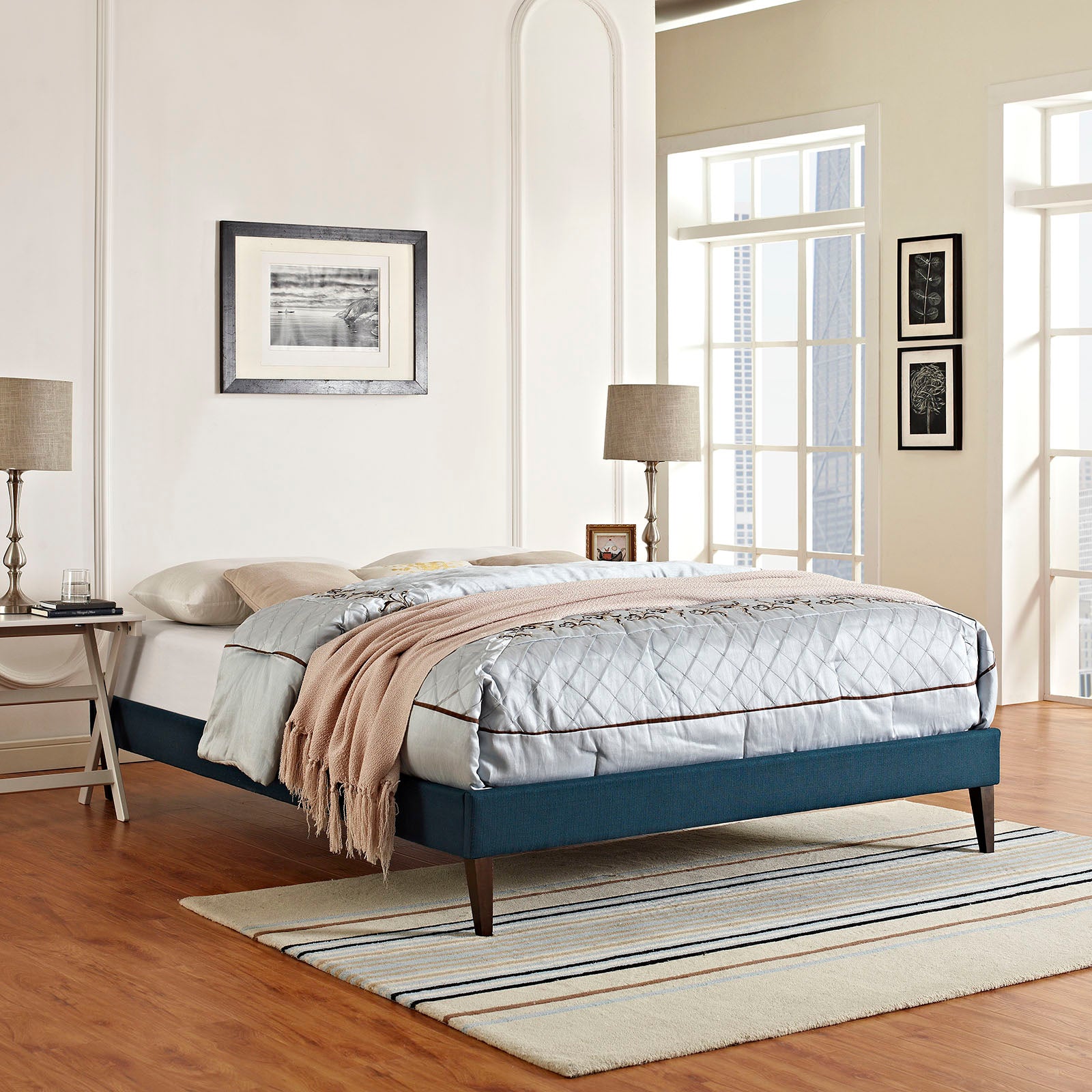 Azure upholstered platform bed with quilted gray comforter, brown piping, beige throw; dark wood legs in a modern bedroom.