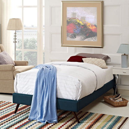 Teal upholstered twin bed frame with white linens and a light blue throw, round splayed legs, beside a multicolor striped rug and a beige chair.