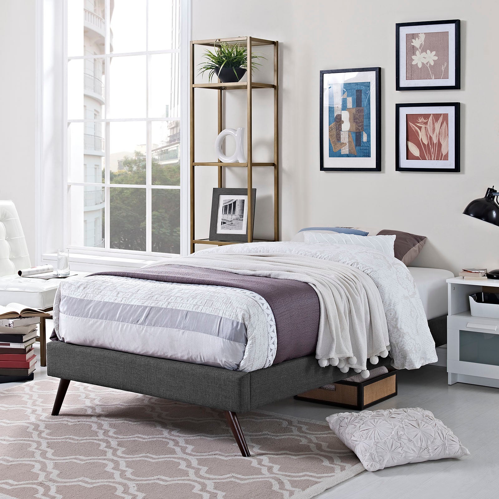 Grey fabric twin bed frame with round splayed legs; mauve-white quilt and white throw, in a bright modern bedroom with a gold shelf unit.