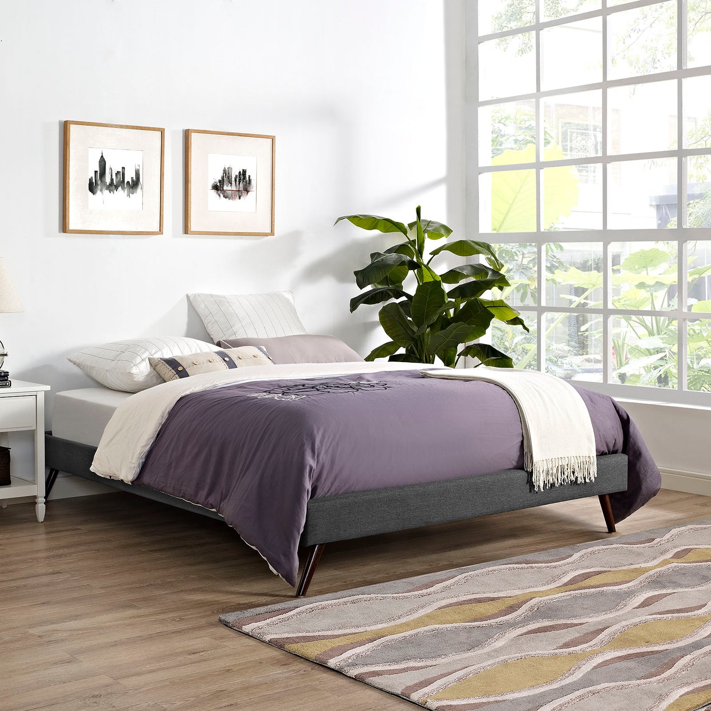 Gray upholstered platform bed with lavender duvet and white throw in a bright modern bedroom; large window, and plant.