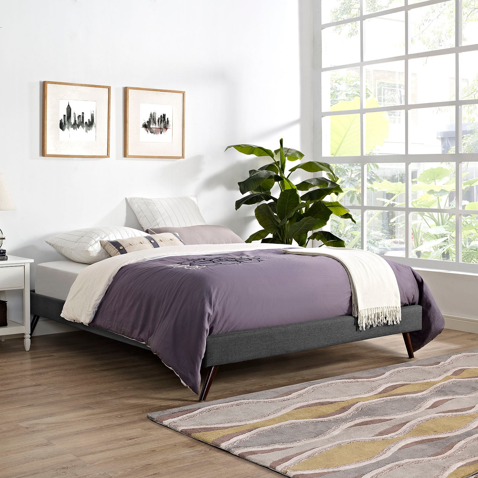 Gray upholstered platform bed with lavender duvet and white throw in a bright modern bedroom; large window, and plant.