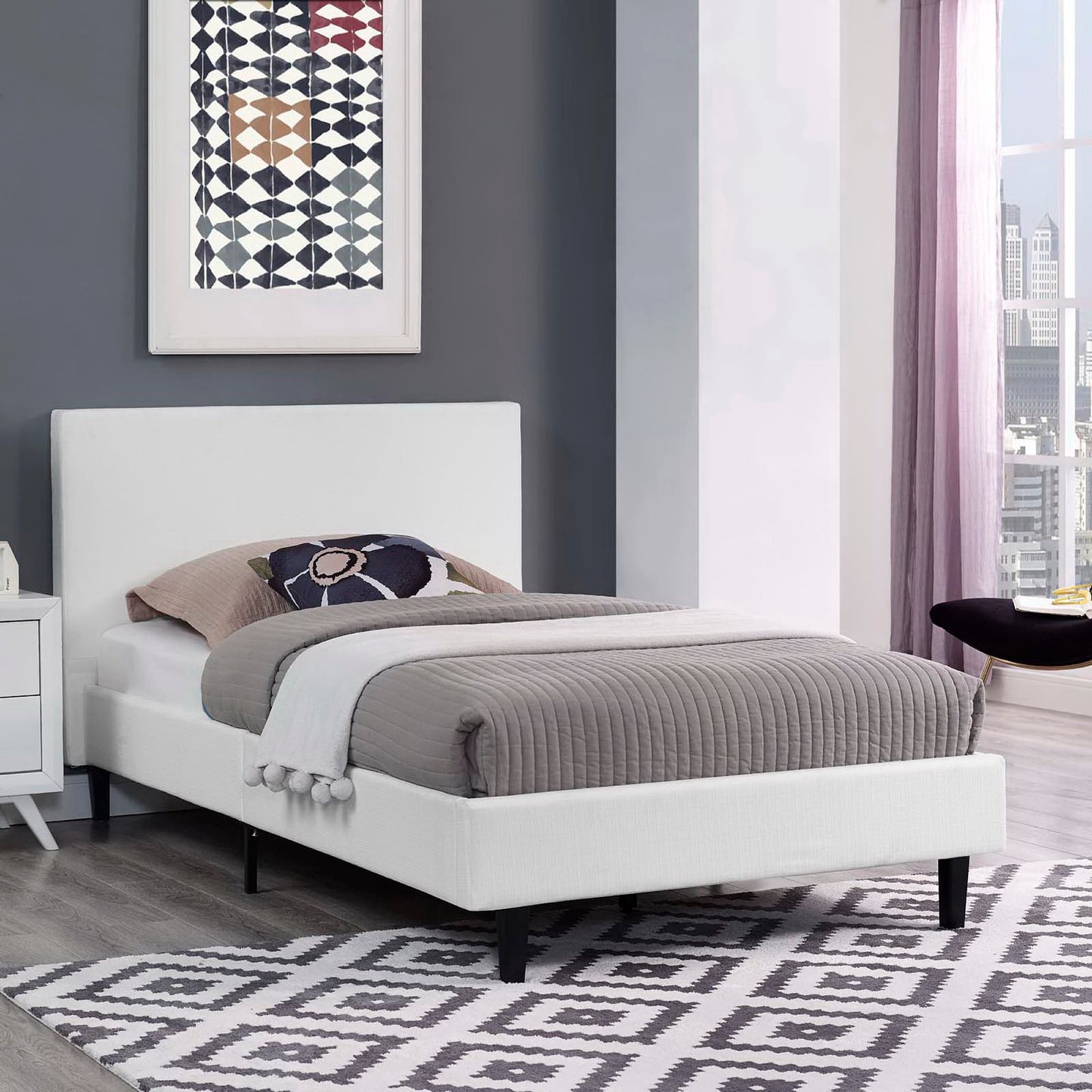 White upholstered platform bed with low headboard, gray quilted cover, beige & navy pillows, black legs, geometric rug.