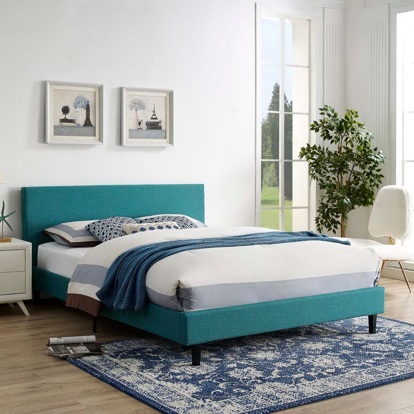 Teal fabric full bed with blue bedding, white nightstand, two framed prints, and a blue patterned rug.