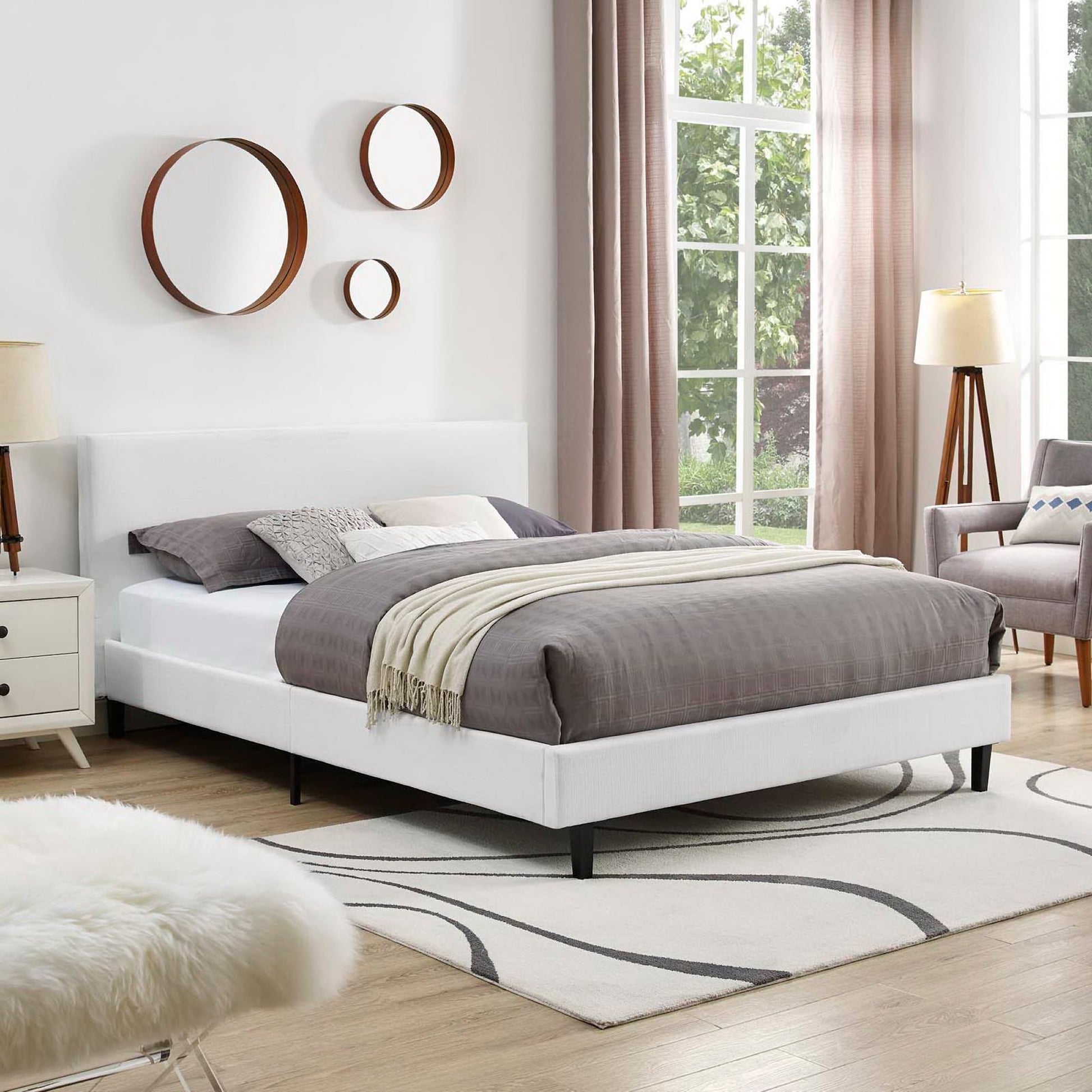 White upholstered platform bed with gray bedding, wood-framed mirrors, light wood nightstand, beige curtains, swirled rug.