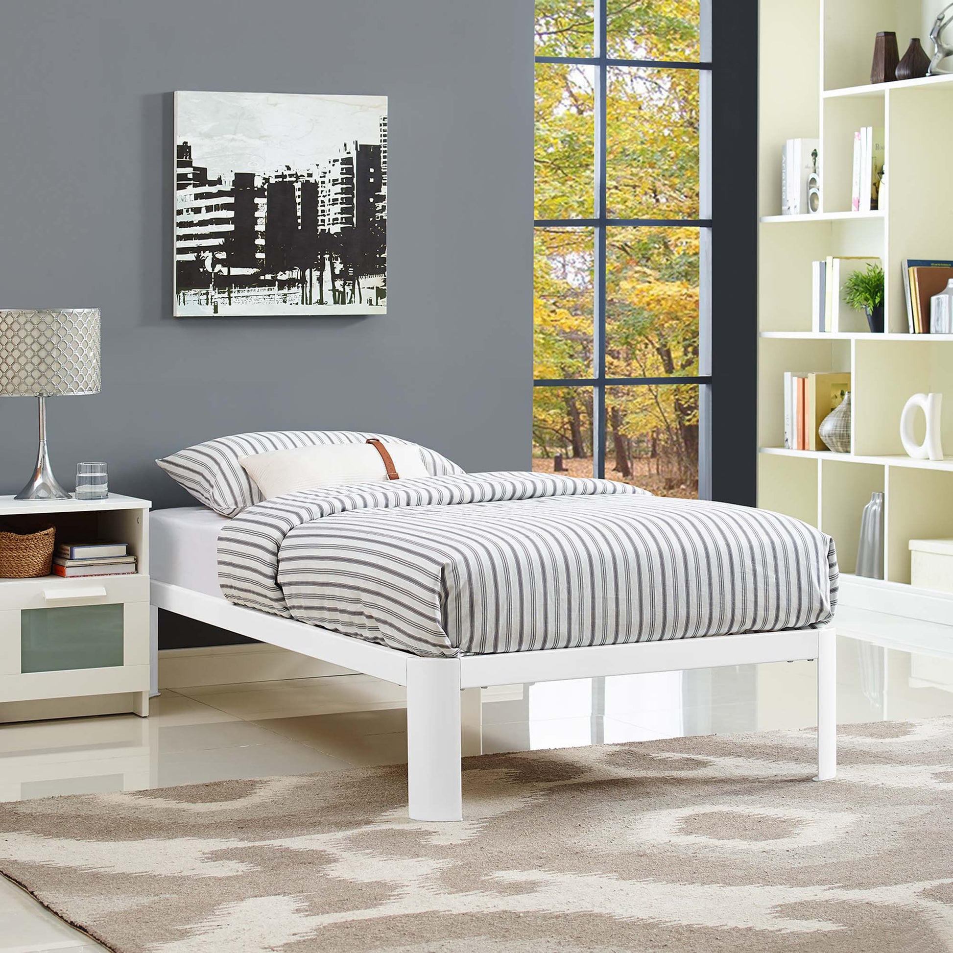 White platform bed with gray striped bedding, gray wall, nightstand lamp, beige rug, and a large window showing fall trees.