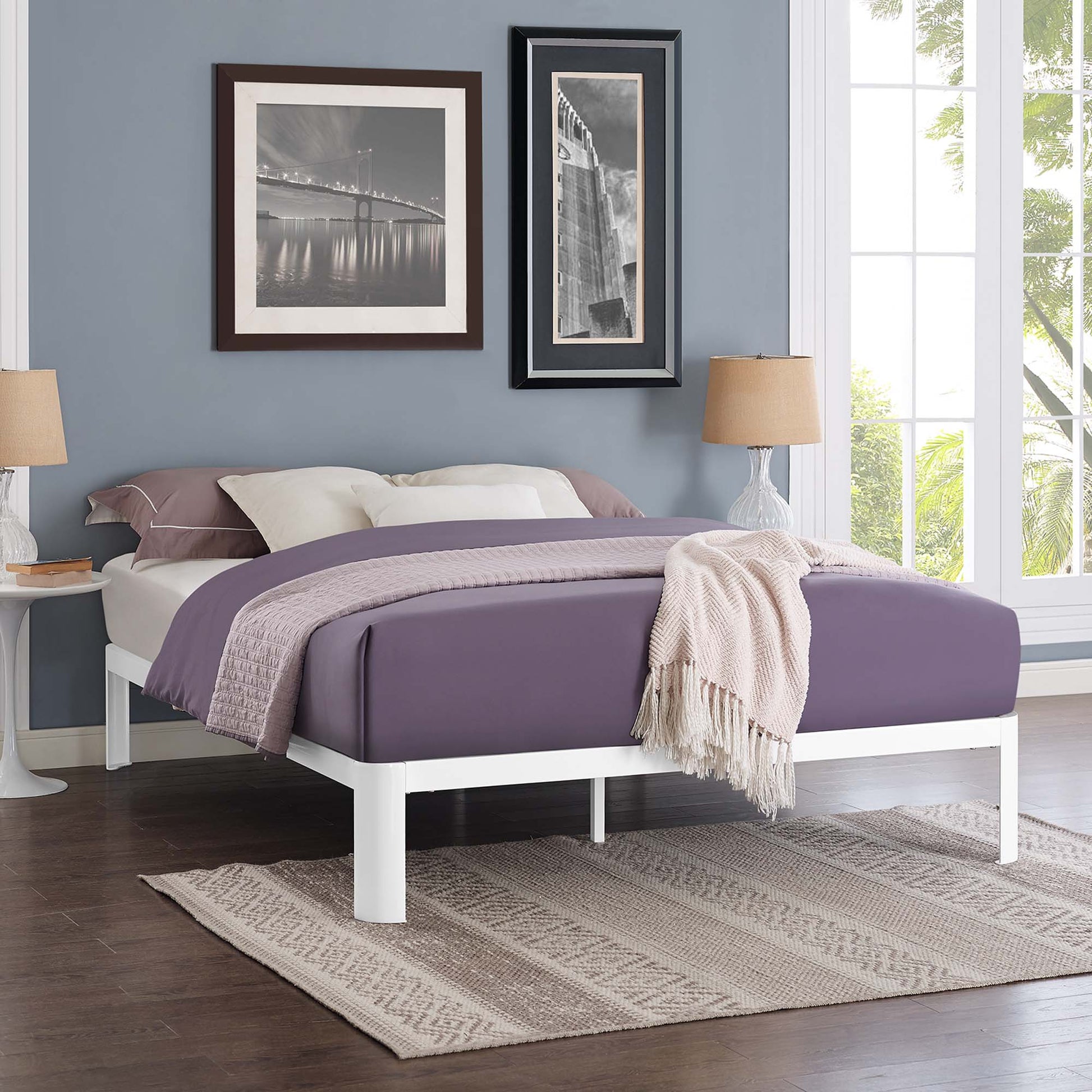 White full bed frame with lavender bedding and a beige throw in a modern blue-walled room with black-framed wall art.