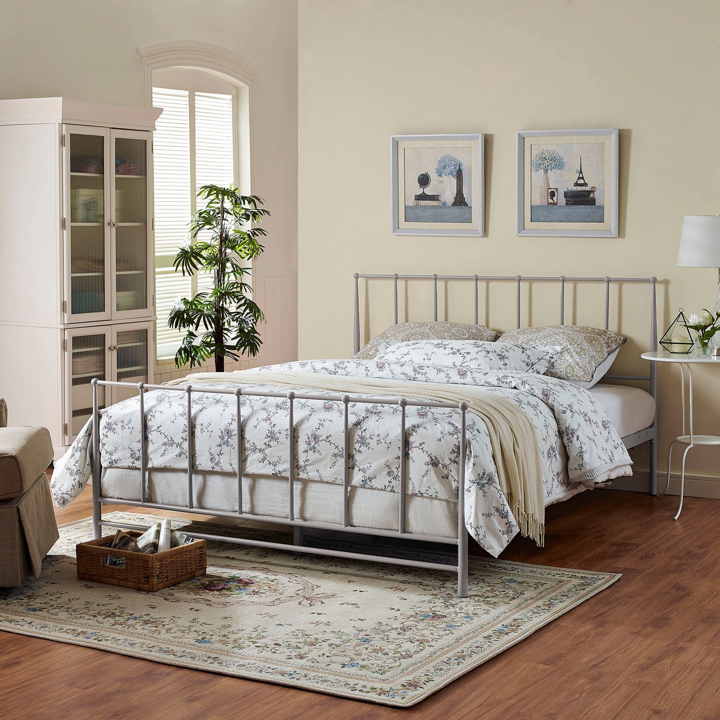 Gray metal bed frame with floral white bedding, beige throw, beige walls, white cabinet, plant, framed art, and floral rug.